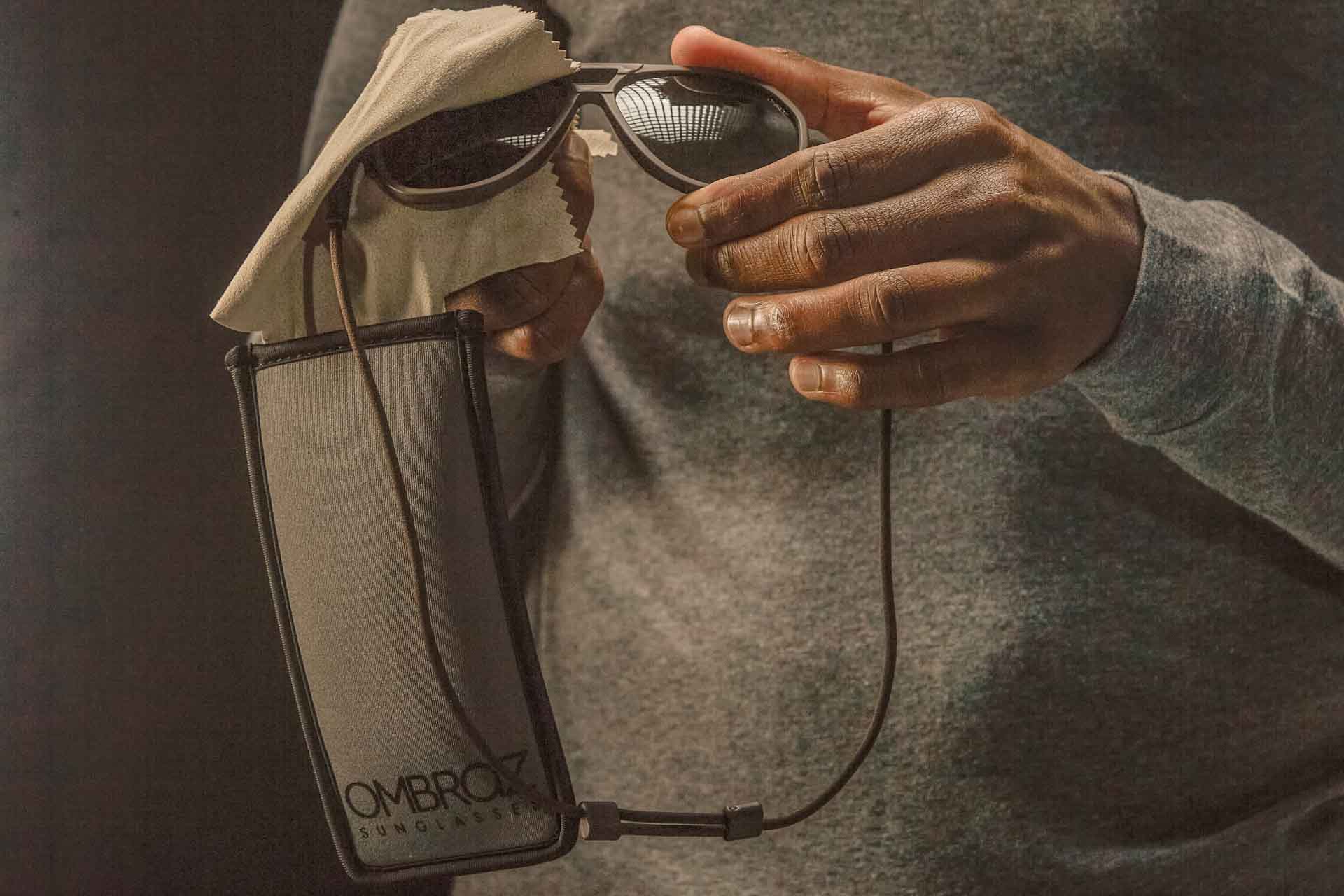 charcoal_grey Hands cleaning a pair of Ombraz classic armless sunglasses with cord using the microfiber cloth in the case