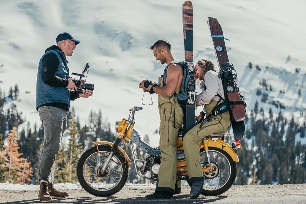 ember_yellow Two skiers on a motorcycle talking to a photographer holding pairs of Ombraz armless rope sunglasses