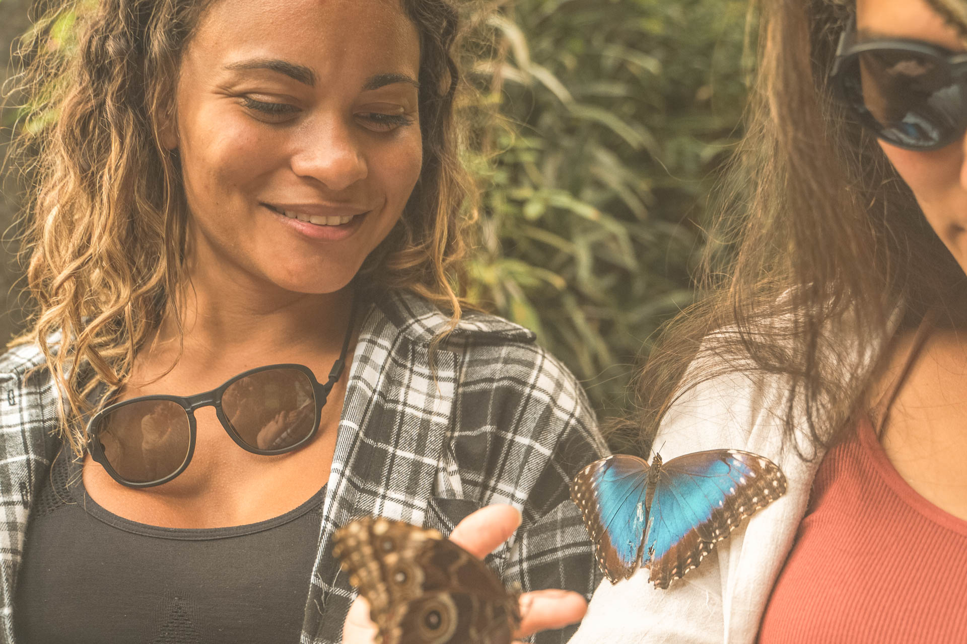 Two women smiling with butterflies carrying Ombraz sunglasses without arms around their necks TETON_CHARCOAL_BROWN