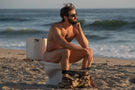 TETON_TORTOISE_GREY Man on a toilet on the beach wearing Ombraz armless sunglasses with cord