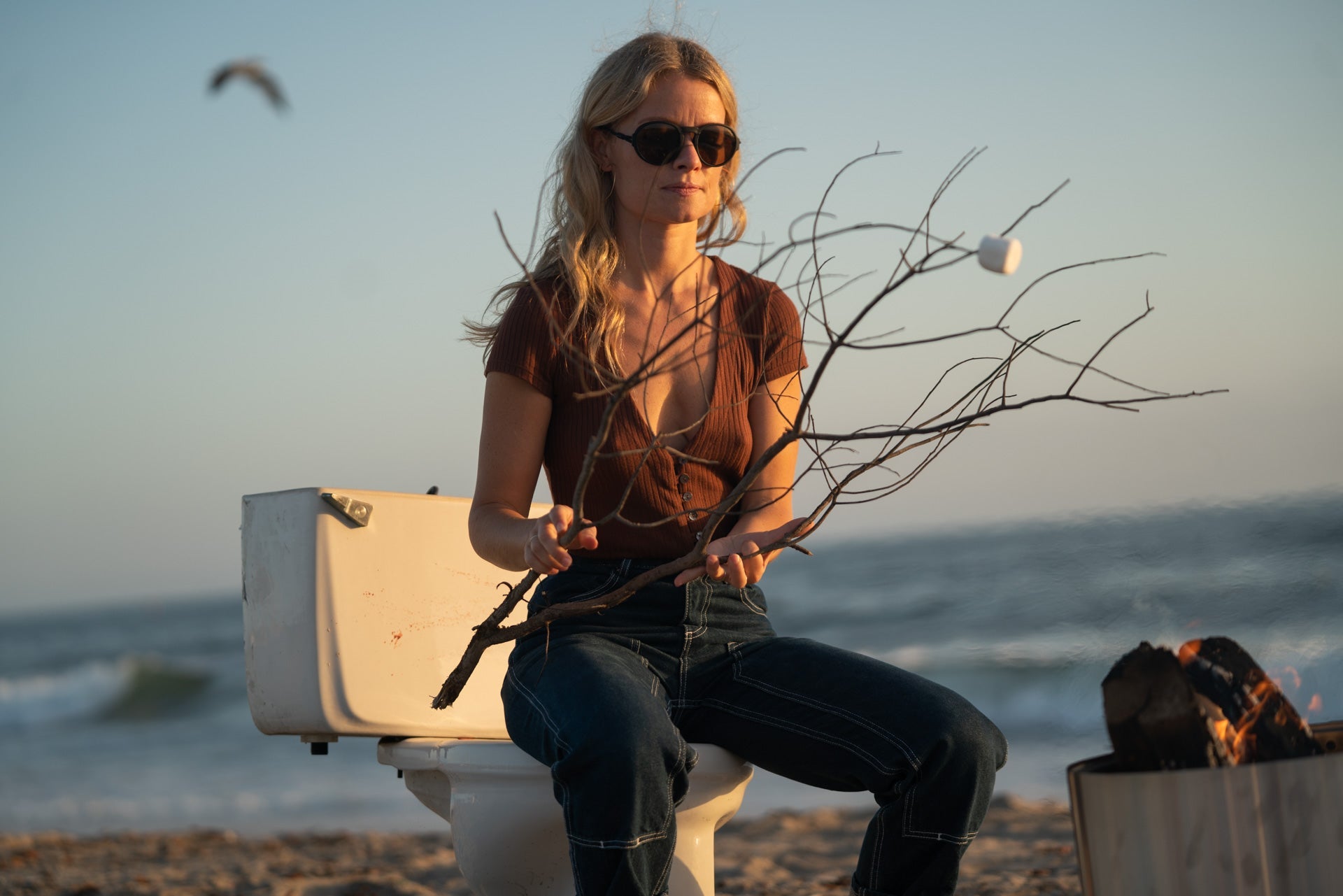 VIALE_CHARCOAL_BROWN Woman at the beach sitting on a toilet roasting marshmallows wearing Ombraz armless strap sunglasses