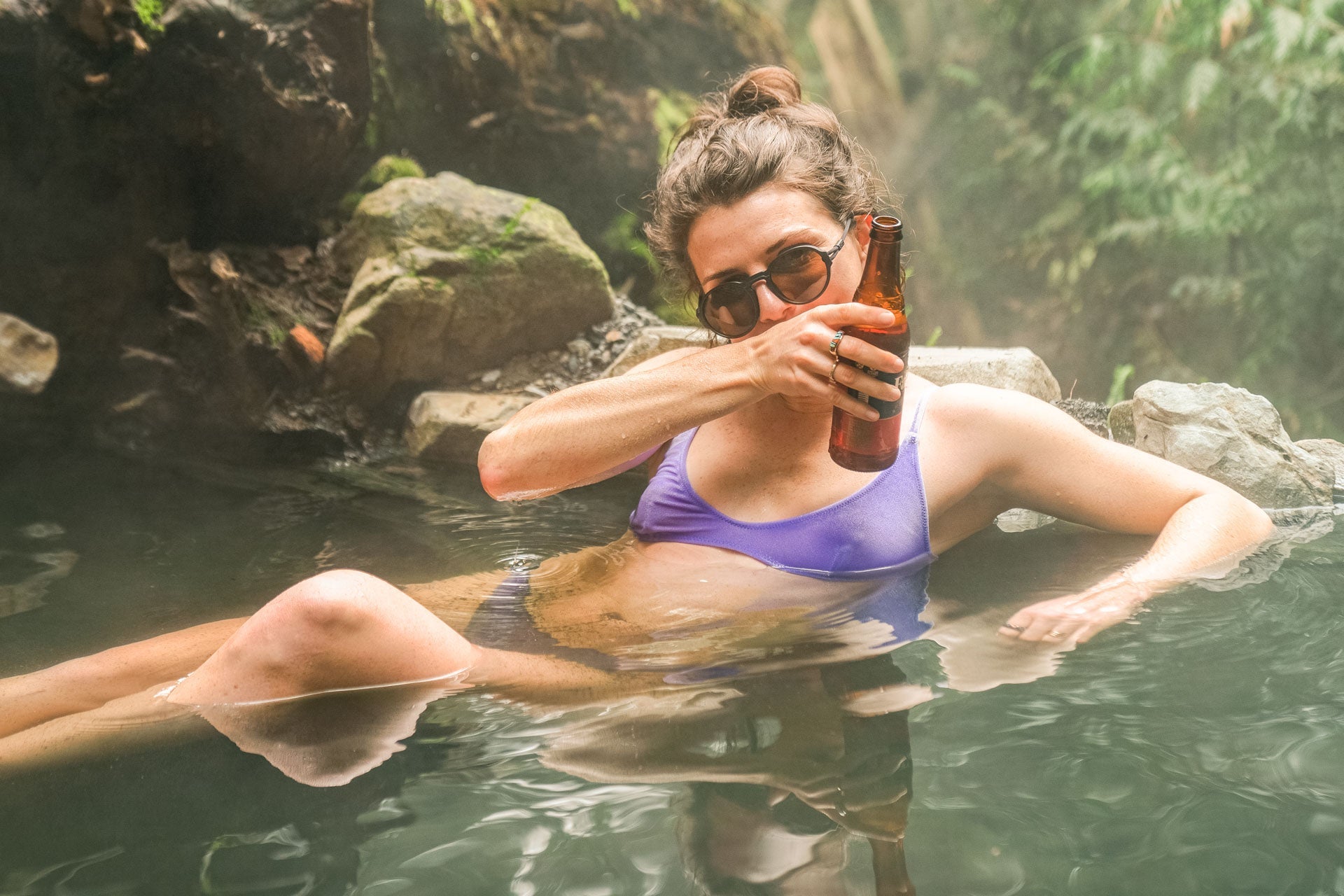 VIALE_CHARCOAL_BROWN Woman drinking a beer in a pond wearing Ombraz armless rope sunglasses