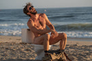 VIALE_CHARCOAL_GREY Man laughing naked on a toilet at the beach wearing Ombraz armless sunglasses with cord