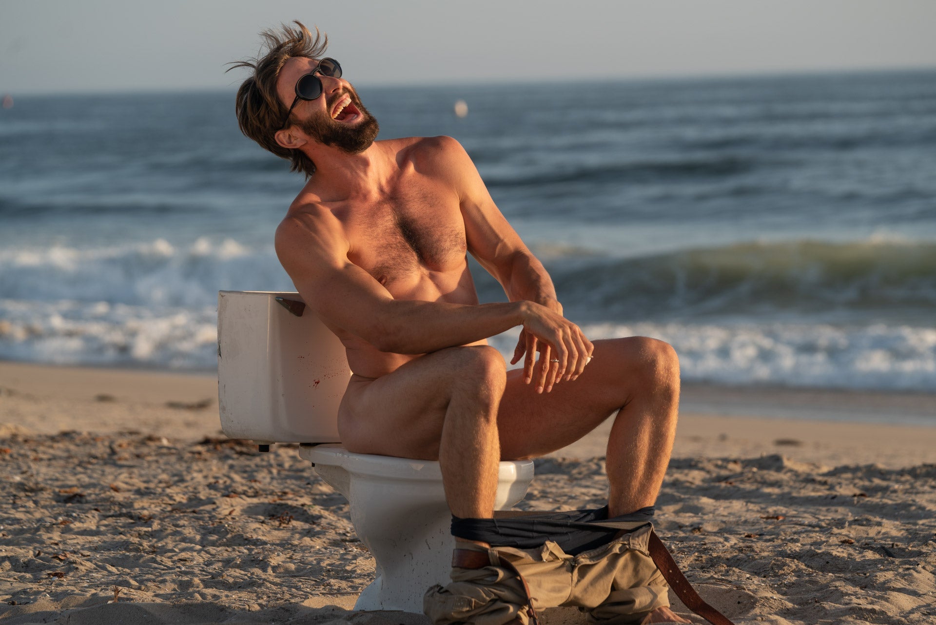 VIALE_CHARCOAL_GREY Man laughing naked on a toilet at the beach wearing Ombraz armless sunglasses with cord